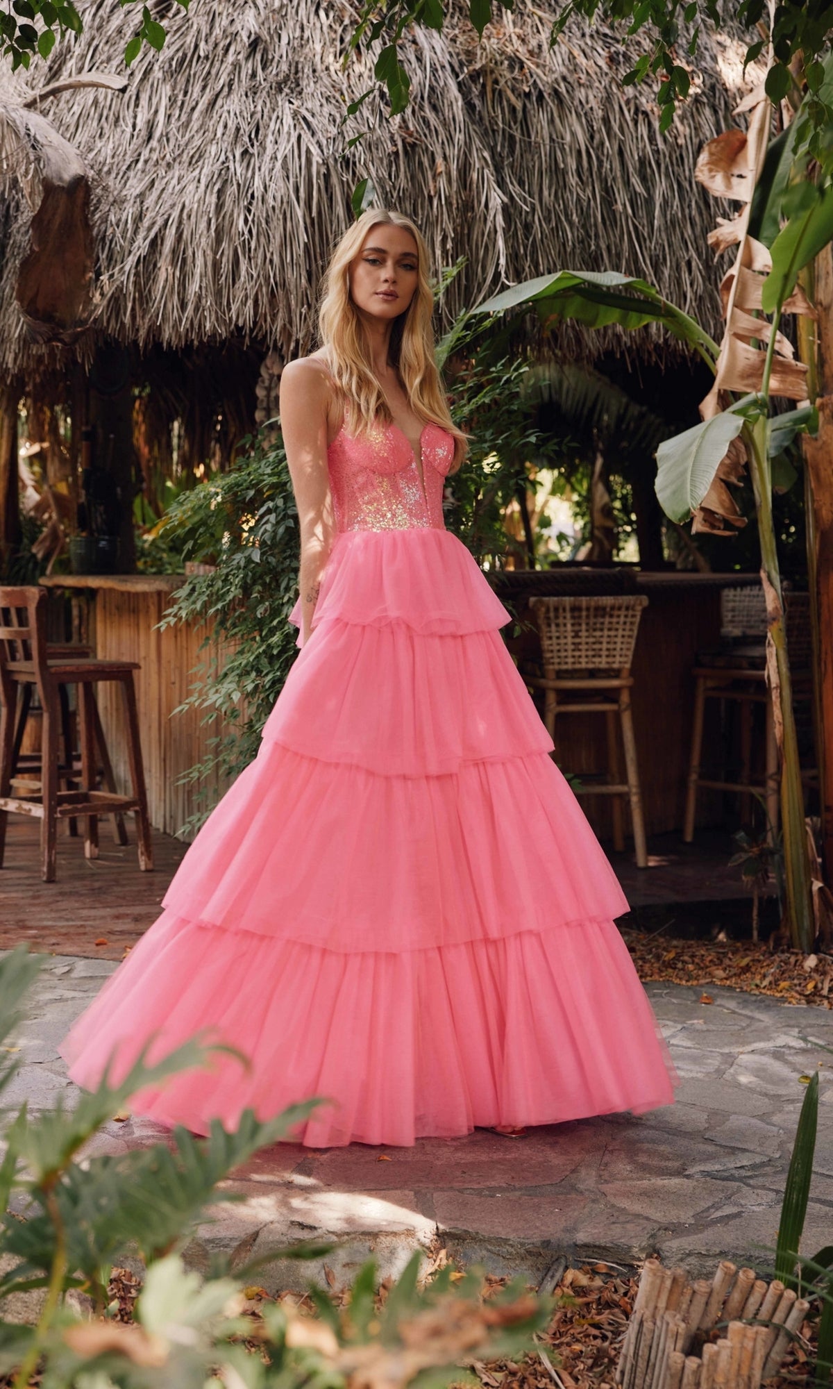 A woman stands outdoors in the Long Prom Dress JT2457H by Juliet, a floor-length ruffle tulle ball gown with a fitted bodice and gold detailing. Thatched roof and bar stools appear in the background.